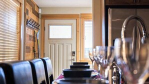 Een eetruimte met een lange tafel gedekt voor vier personen, een raam met jaloezieën, en een keuken met een koelkast en kasten.