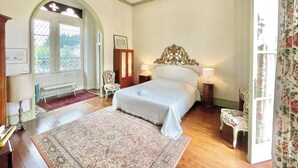 A bedroom with a large bed, a wooden arched doorway, a chair, a nightstand, and a window with a view of greenery. - Manoir de Cénac 19th century - with swimming pool and large park (Cénac-et-Saint-Julien)