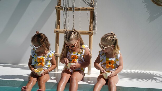 Tres nens asseguts al costat d'una piscina, portant gandules de bany amb un dibuix de fruita.