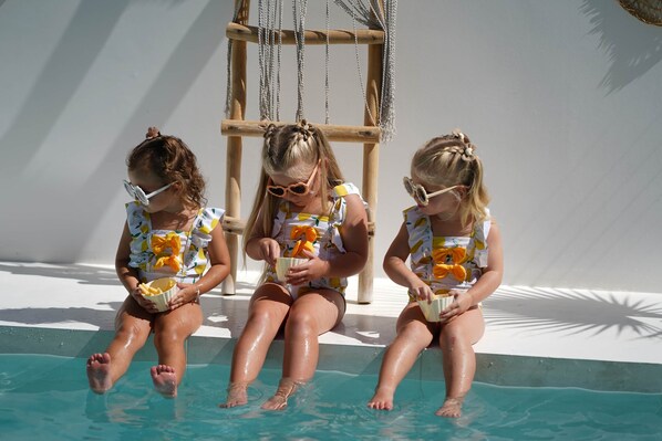 Drei Kinder sitzen am Pool, sie tragen passende Badeanzüge mit Obstmuster.