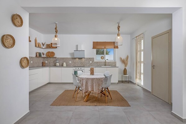 A modern kitchen with white cabinets, a granite countertop, and a dining area with a round table and chairs. - Villa-Pastida (Rhodes)