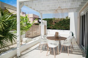 Eine überdachte Terrasse im Freien mit einem runden Tisch und weißen Stühlen, umgeben von Pflanzen und Blick auf eine Wohngegend.