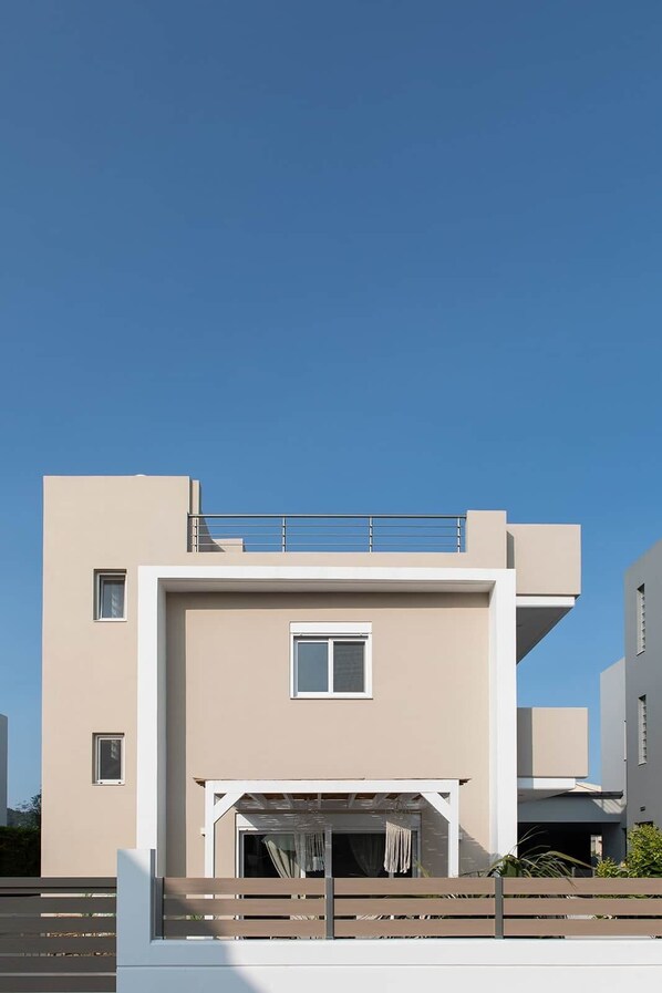 A modern building with a flat roof, a balcony, and a covered entrance with a bench. - Villa-Pastida (Rhodes)