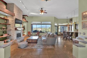A spacious living room with a fireplace, a large window offering a view of the mountains, and a modern kitchen with bar seating. - Casa De Kokopelli - Luxurious Desert Retreat with Hot Tub, Infinity Pool, Spe... (Cave Creek)