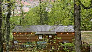 A rustic cabin with a deck and outdoor seating area, surrounded by dense forest.
