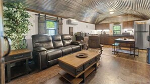 A spacious living area with a leather sofa, a wooden coffee table, and a dining table. The room features a high ceiling with exposed ductwork and a large window providing natural light.