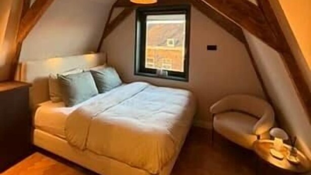 A bedroom with a sloped ceiling, a bed, a window, and a wooden beam structure. - Vermeer - luxury authentic room at Museumplein (Amsterdam)