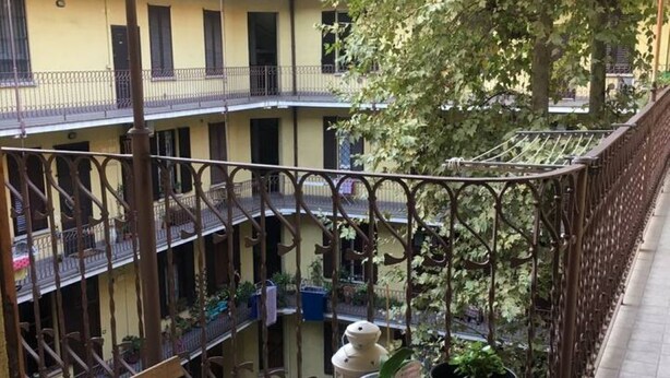 A balcony with a wooden bench, potted plants, and a metal railing overlooking a courtyard with buildings and trees. - Cozy and bright apartment ideal for two people (Milano)