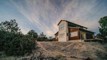 A modern two-story house with a large glass window on the ground floor, surrounded by natural landscape.