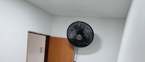 Una habitación pequeña y poco amueblada con una cama individual, un ventilador de pie y una puerta que da a otra habitación.
