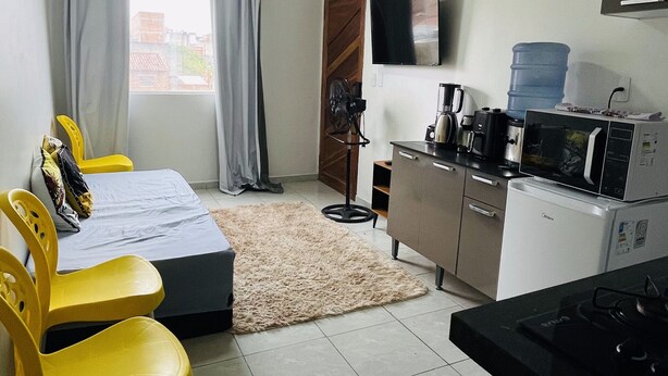 Habitación de hotel compacta con cama, escritorio, silla, kitchenette pequeña, televisor y ventana con cortinas.