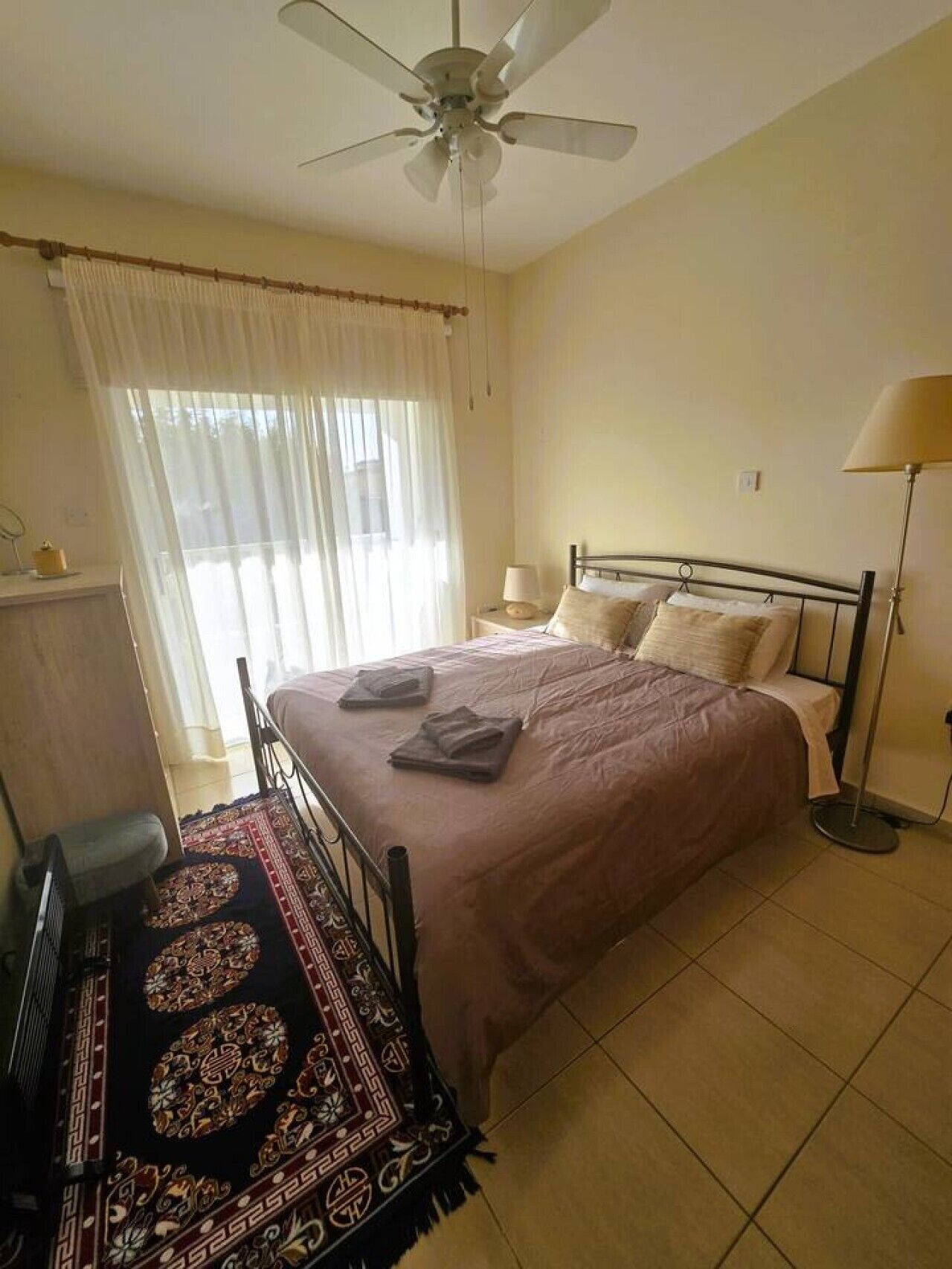 Een slaapkamer met een bed, een plafondventilator, een bedlamp, een vloerlamp en een raam met gordijnen.