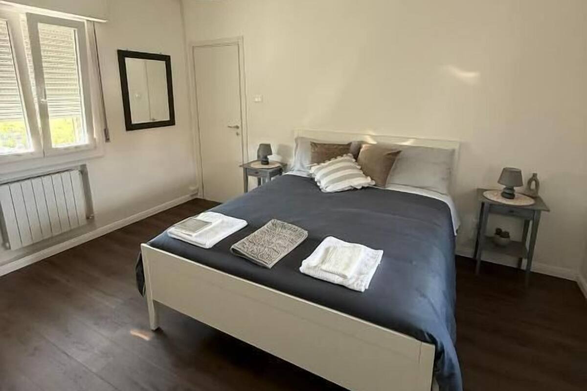 Un dormitorio con cama, mesitas de noche, lámpara colgante, ventana con persianas y radiador.