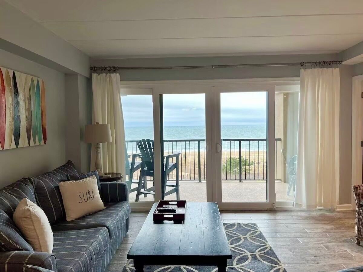 Een woonkamer met een bank, een bijzettafeltje en uitzicht op het strand door grote glazen deuren.