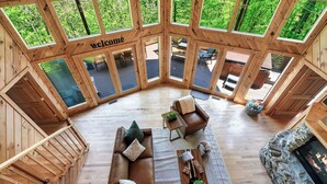 Een ruime houten hut met een 'welcome' bord, een open haard en een gezellige zithoek met een bank en stoelen.
