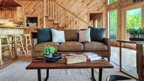 Een gezellig interieur van een chalet met een houten trap, een bruine leren bank met decoratieve kussens, een salontafel met boeken en een plant, en uitzicht op groen buiten.