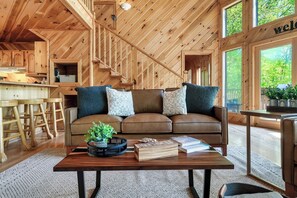 Ein gemütliches Blockhaus-Interieur mit einer Holztreppe, einem braunen Ledersofa mit gemusterten Kissen, einem Couchtisch mit Büchern und einer Pflanze sowie Blick ins Grüne.