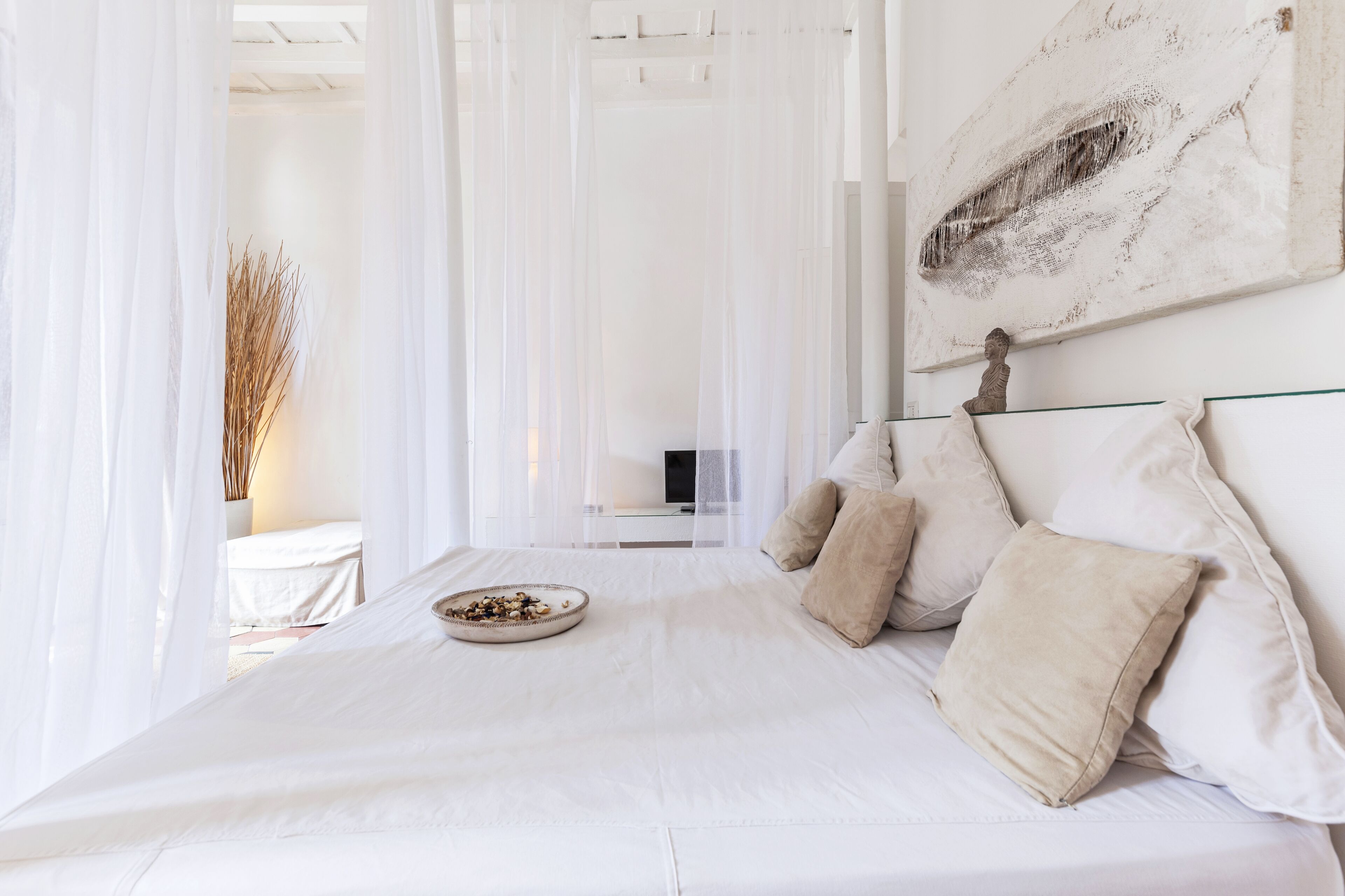 A large bed with white linens, a bowl of fruits, and beige pillows.
