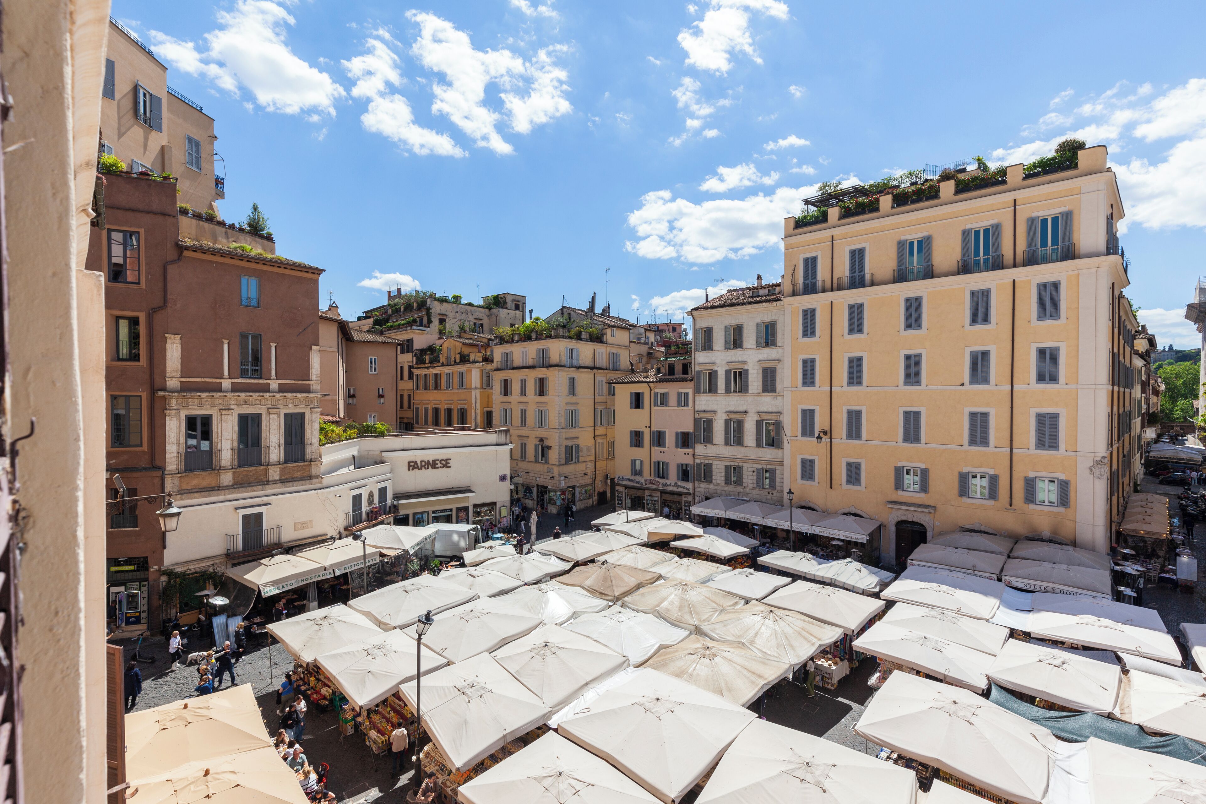 A bustling outdoor market area with numerous white umbrellas, surrounded by multi-story buildings with balconies and commercial establishments.