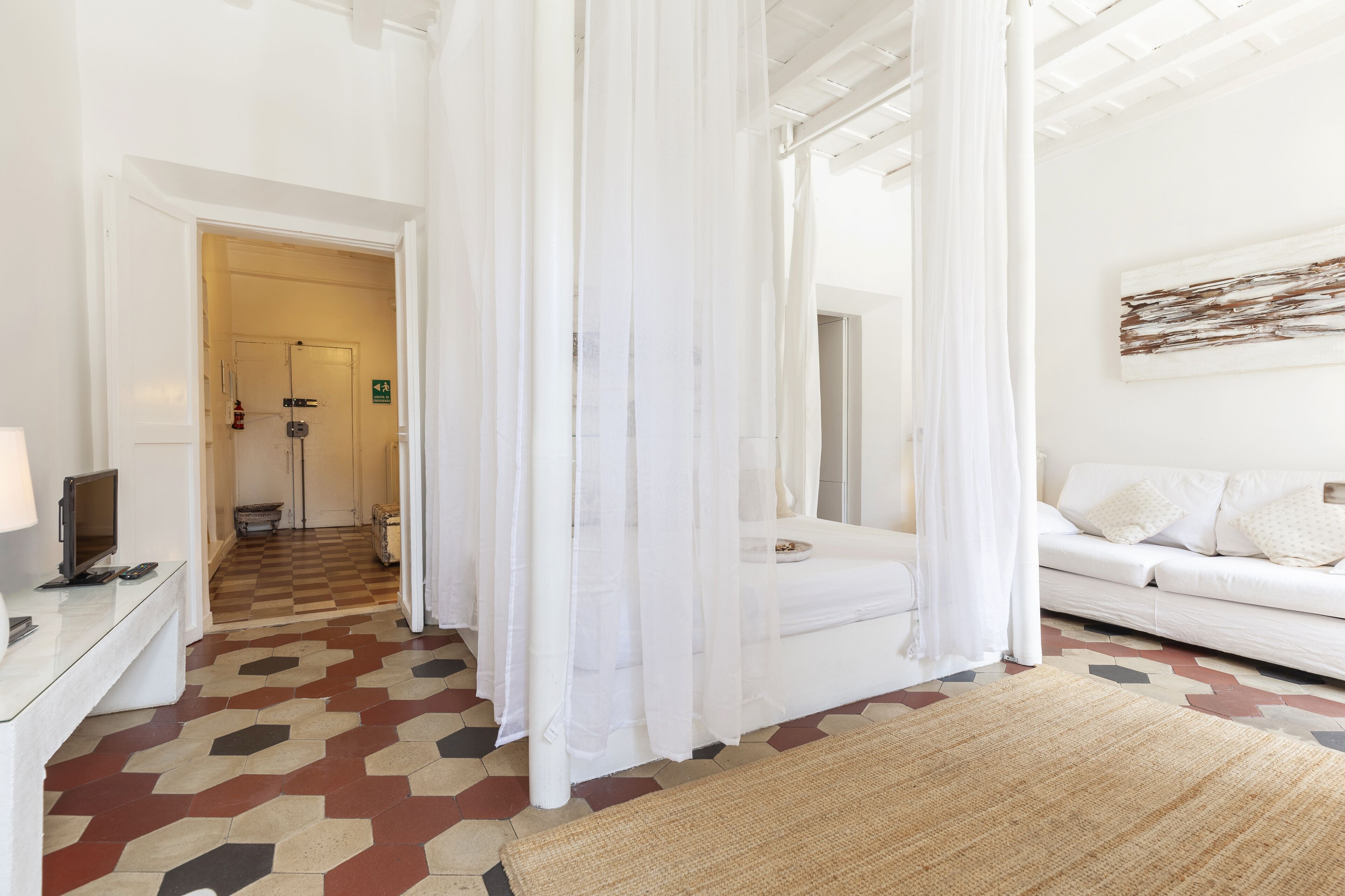 Une chambre d’hôtel avec un sol à carreaux, un canapé blanc, un téléviseur posé sur un bureau et un lit surmonté d’un baldaquin blanc.