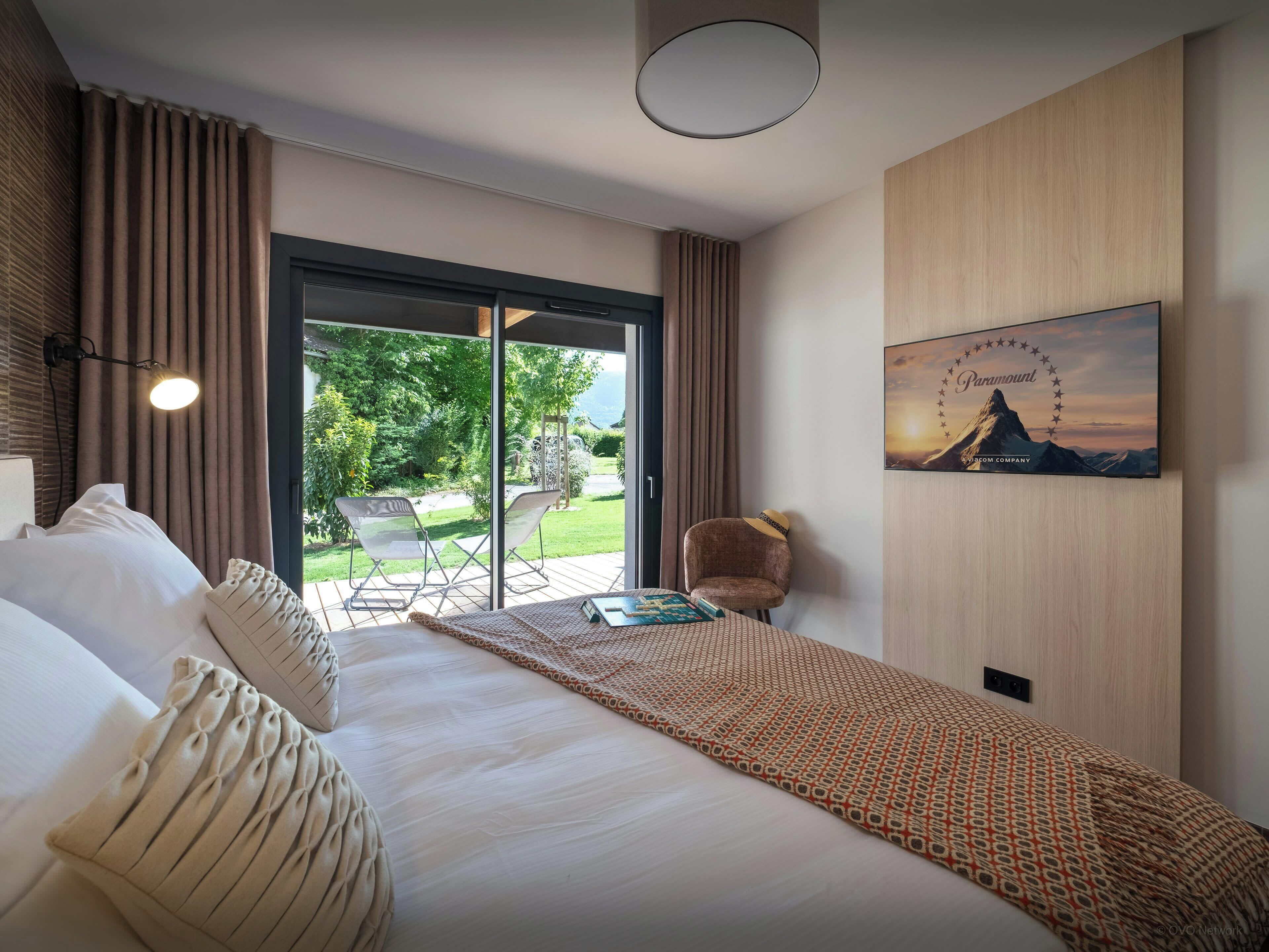 Quarto de hotel com cama, cadeira, mesa, uma janela grande e um quadro na parede.