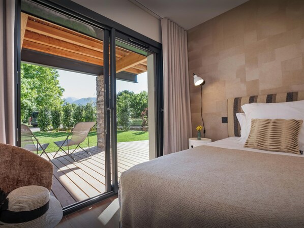 A bedroom with a large bed, a nightstand, a lamp, a window with curtains, and a view of the outdoors.