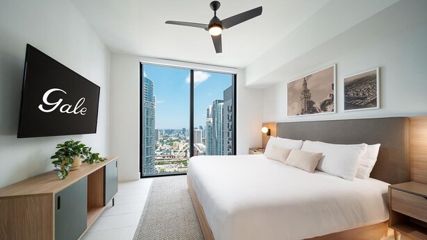 A modern hotel room with a large bed, a ceiling fan, a TV, a city view, and artwork on the walls. - Skyline Residences One Bedroom with Kitchen & Balcony (Miami)