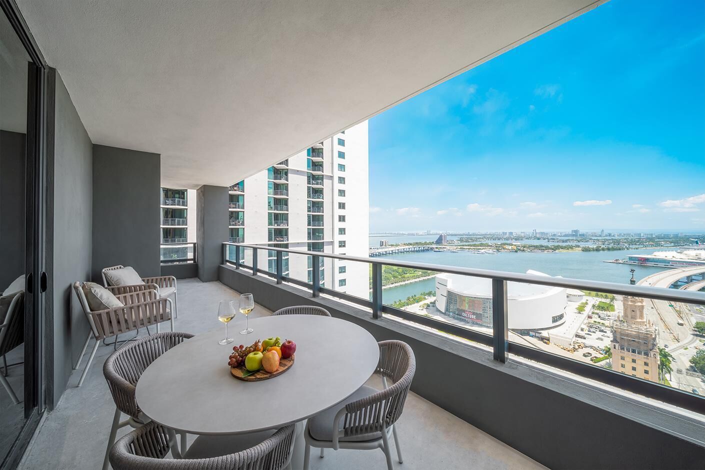 Een balkon met een ronde tafel, rieten stoelen en een schaal fruit met wijnglazen.