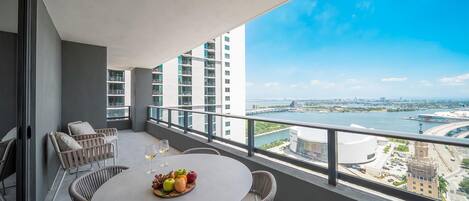 Een balkon met een ronde tafel, rieten stoelen en een schaal fruit met wijnglazen.