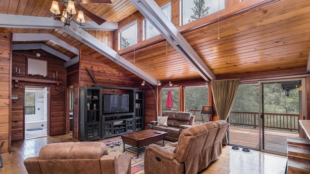 A spacious living room with a large TV, comfortable seating, and a balcony with a view of the forest.