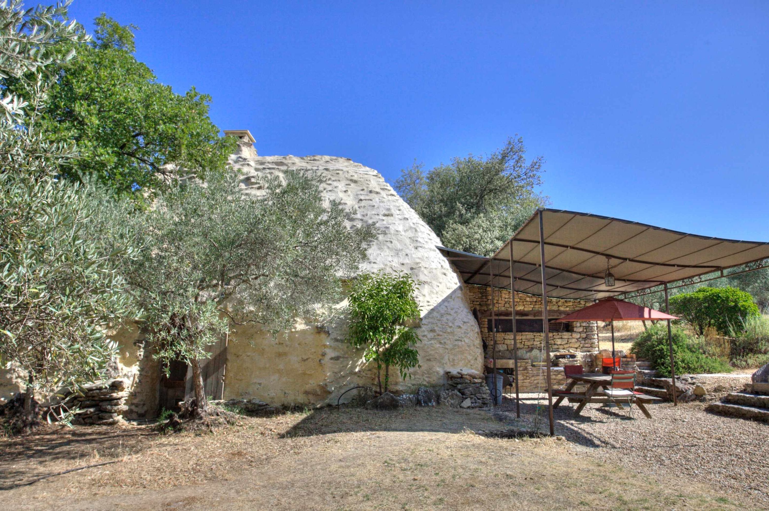 A rustic outdoor area with a stone wall, olive trees, and a covered patio with a table and chairs.