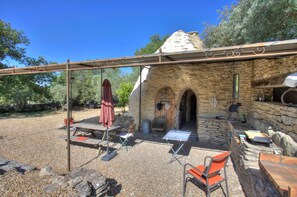 Un bâtiment rustique en pierre, doté d’une porte en bois et d’une petite fenêtre, entouré de gravier et d’arbres.
