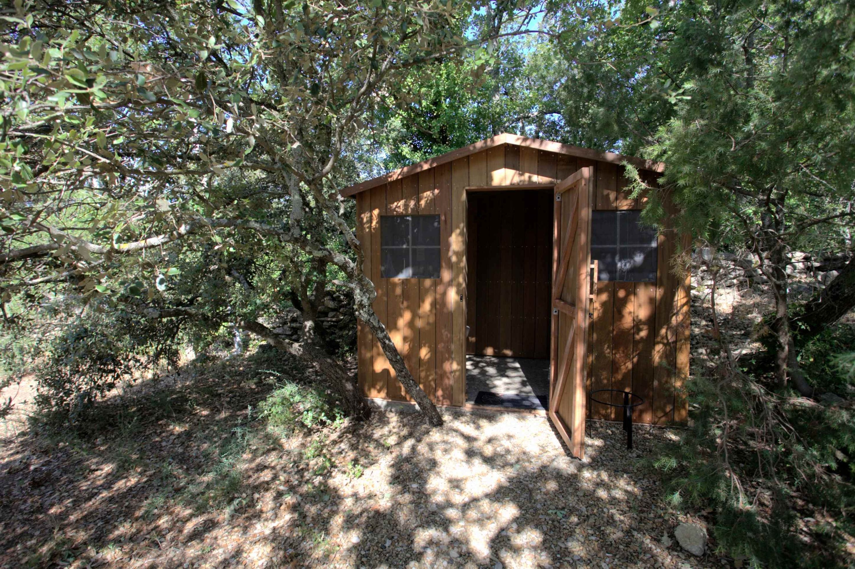 A small wooden cabin with a door open, surrounded by trees and natural ground cover.