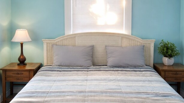 Een bed met een gestreept dekbedovertrek, twee kussens, een nachtkastje met een lamp, een potplant en een raam met jaloezieën.