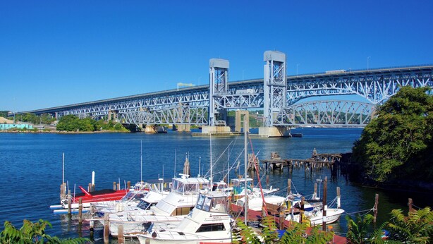 A marina with boats docked, a large blue bridge in the background, and clear blue skies.