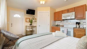 A bedroom with a bed, a chair, a desk, and a kitchen area with a microwave and stove.