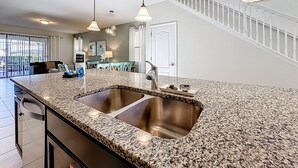 Uma cozinha com bancadas de granito, pias de aço inoxidável, iluminação pendente, uma escadaria e uma área de jantar com cadeiras e mesa.