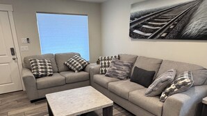 A modern living room with a sectional sofa, a coffee table, patterned throw pillows, a large window, and a wall-mounted artwork.