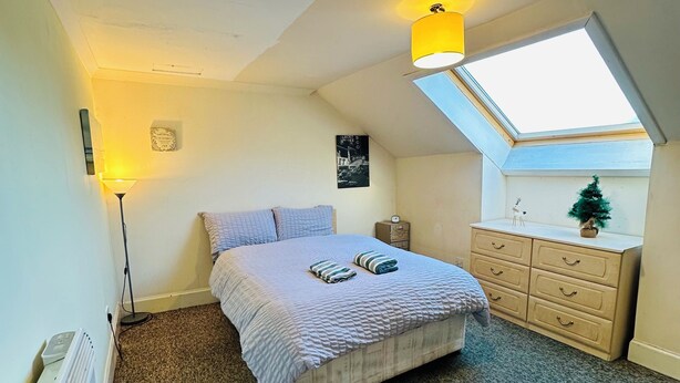 Une chambre à coucher comprenant un lit, une commode, un radiateur, une lampe, une petite décoration en forme d’arbre et une lucarne.
