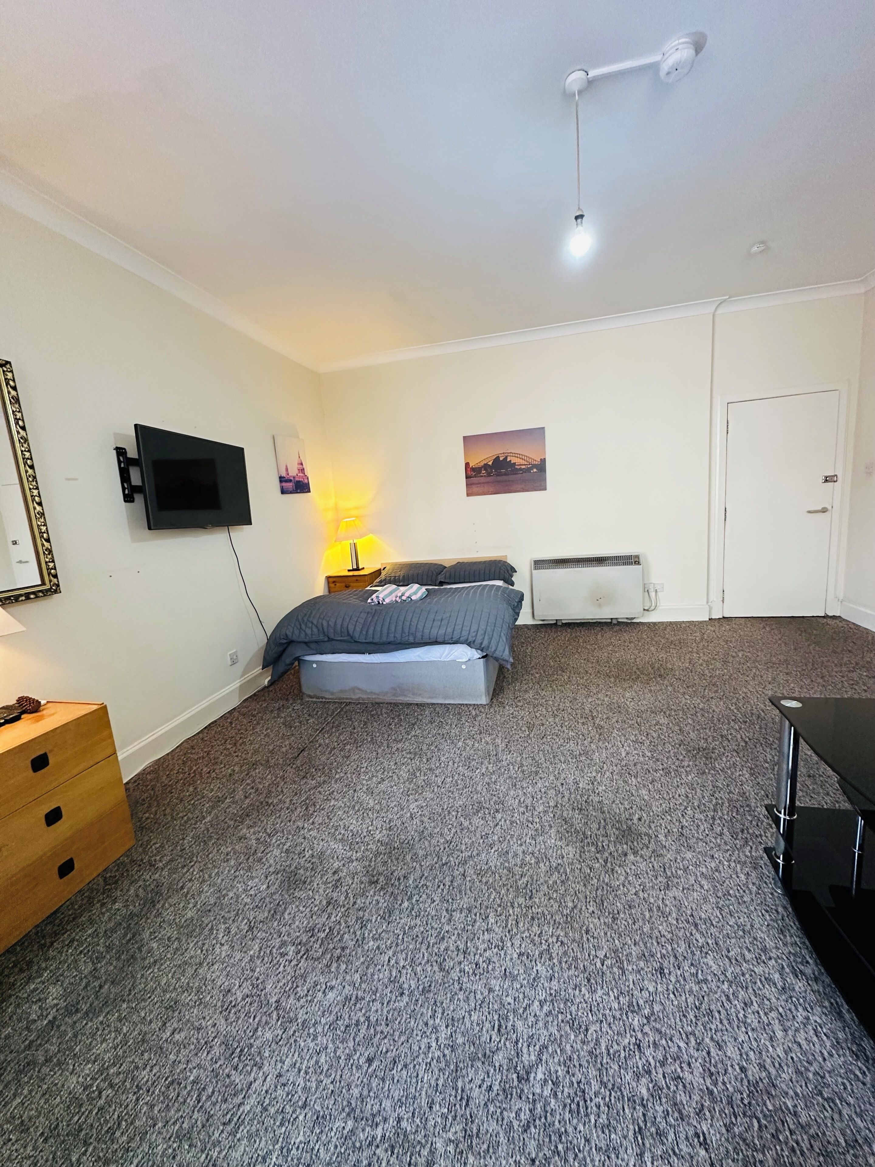 Un dormitorio con cama, televisor en la pared, lámpara, calentador y puerta.