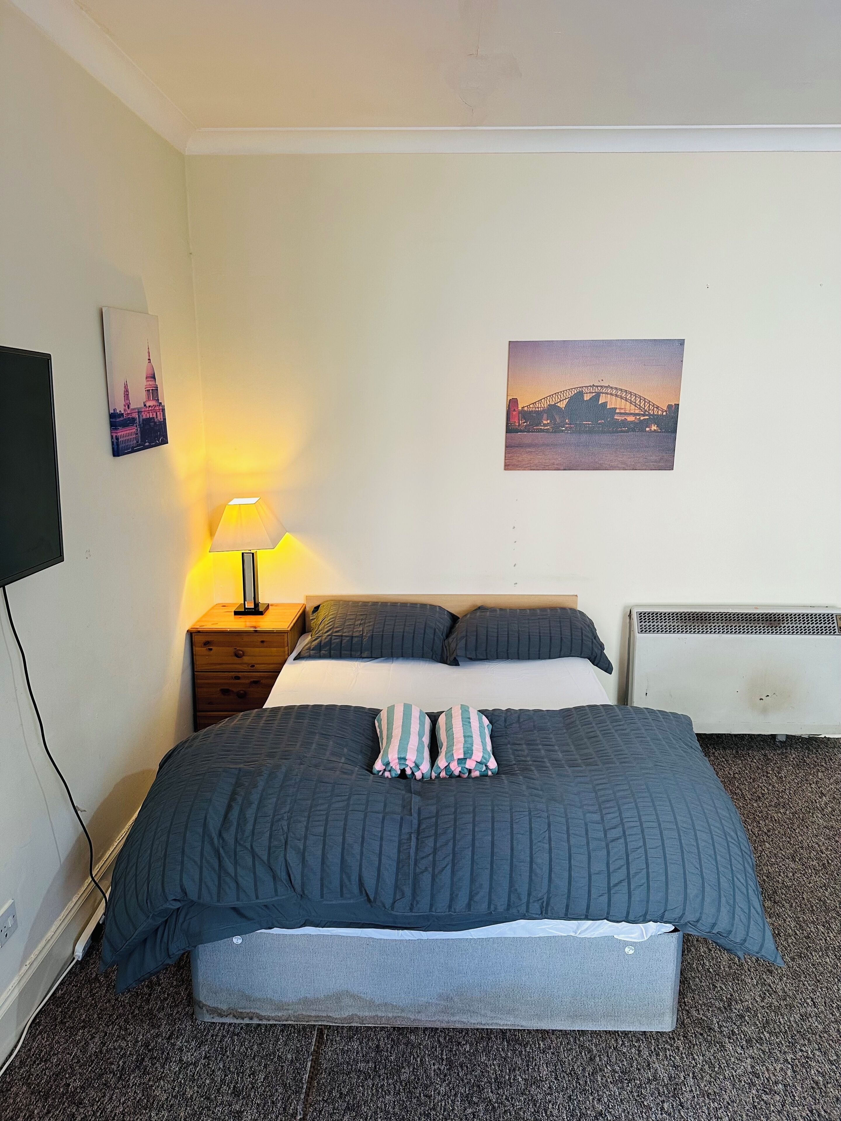 A bedroom with a bed, a bedside lamp, a heater, and two wall-mounted pictures.