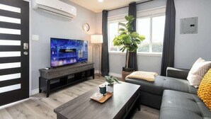 Ein modernes Wohnzimmer mit Flachbildfernseher, dunklem Sofa, Couchtisch und großem Fenster mit Vorhängen.