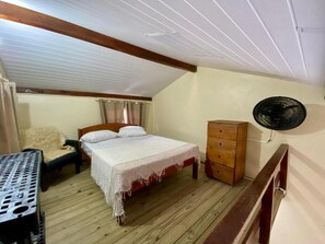 Un dormitorio con piso de madera, una cama con cubrecama blanco, un aparador de madera y un ventilador de techo.