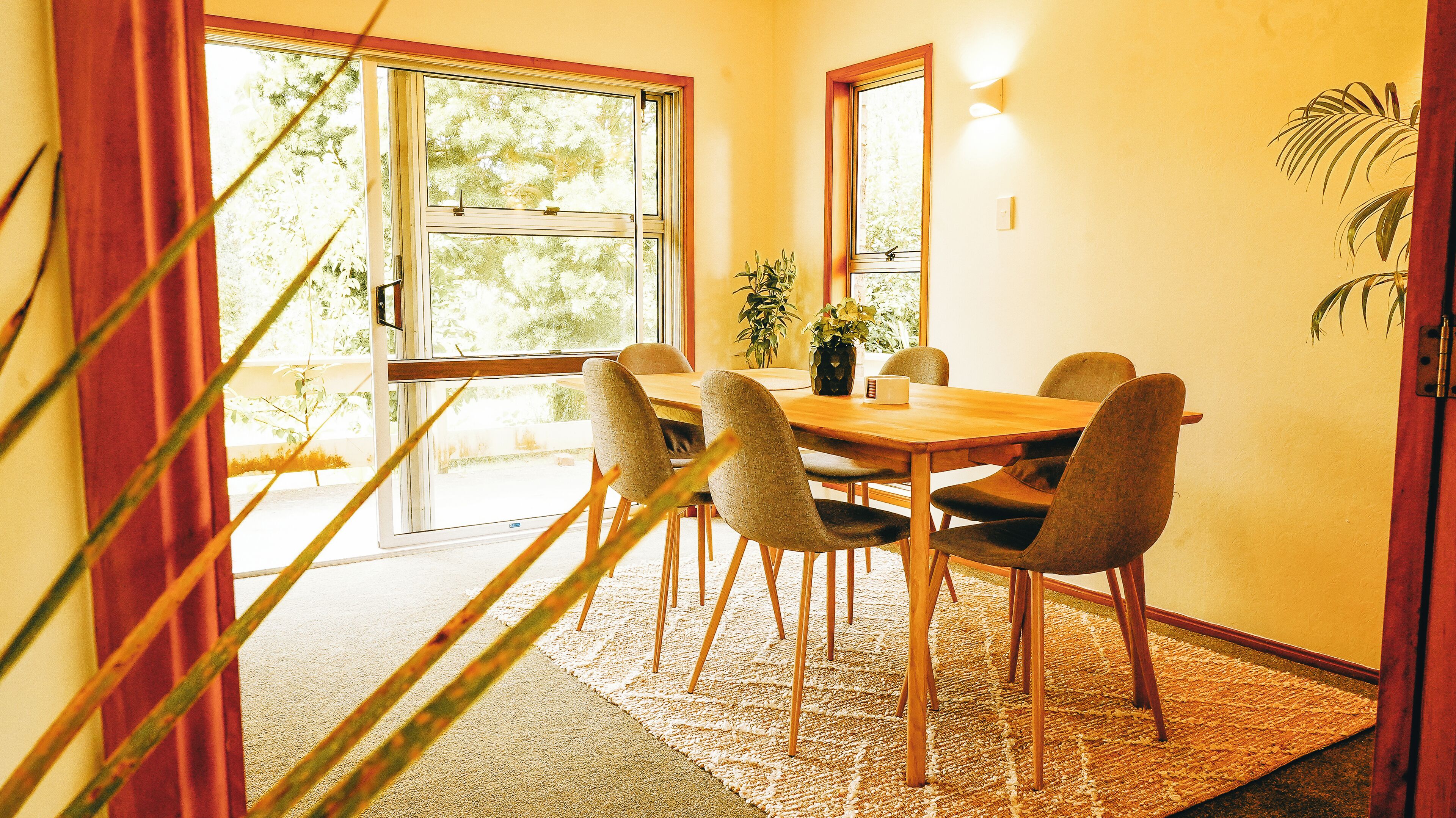 A dining area with a wooden table and chairs, a potted plant, and a view through a glass door.