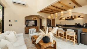 A modern living room with a sofa, a coffee table, and a TV. There is a kitchen area with a counter and bar stools. - Luxe Villa 10p, Grill, 2 Private Pools w/Concierge (Tulum)
