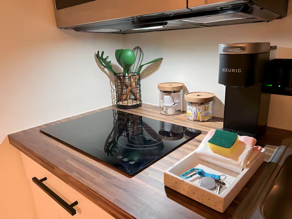 Et køkken med en sort induktionskogetop, en Keurig kaffemaskine, en lille plante og en bakke med køkkenredskaber.