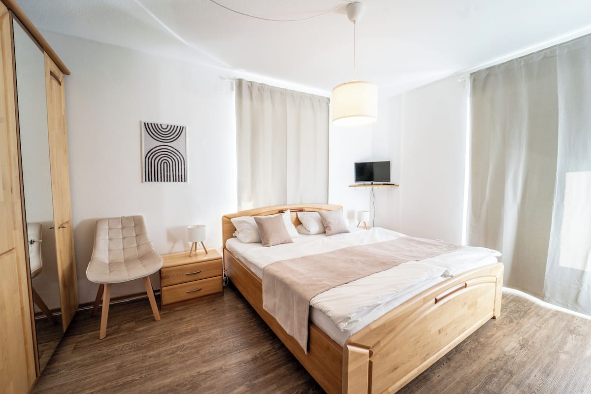 Une chambre moderne avec un lit en bois, une chaise, une table de chevet et un téléviseur fixé au mur.