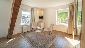 Une pièce avec une poutre en bois au plafond, deux fenêtres avec des rideaux, une petite table avec des chaises et un tapis à motifs.