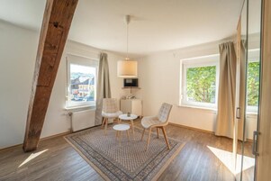 A room with a wooden ceiling beam, two windows with curtains, a small table with chairs, and a patterned rug.