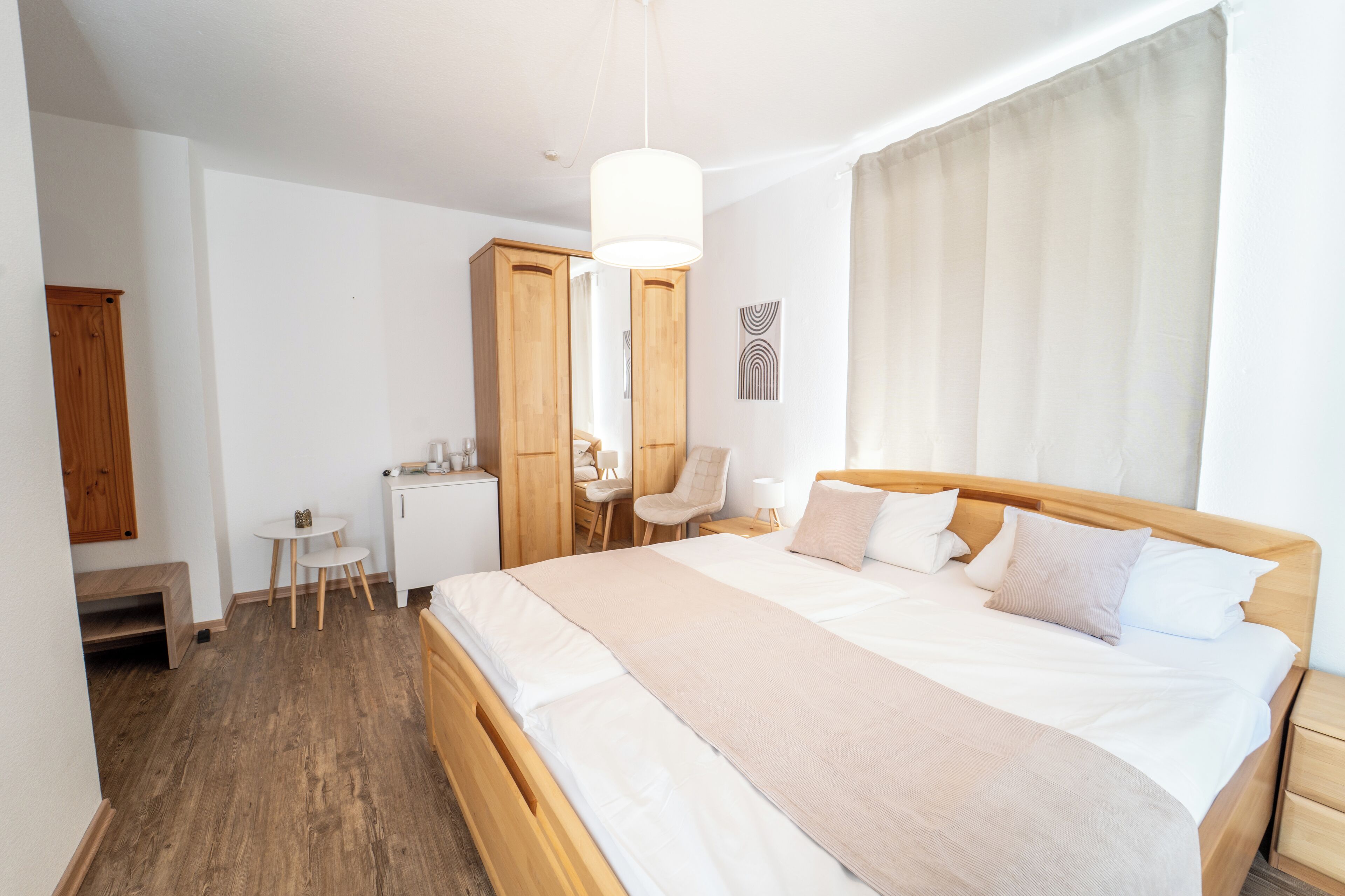A modern bedroom with a large bed, wooden headboard, a bedside table, a small desk, a chair, and a hanging light fixture.